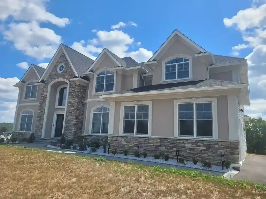 Large luxury two-story home with tan stucco exterior, stone facade entryway tower and base surround, and white decorative trim by Plaster Pro Stucco Contractors serving North Jersey